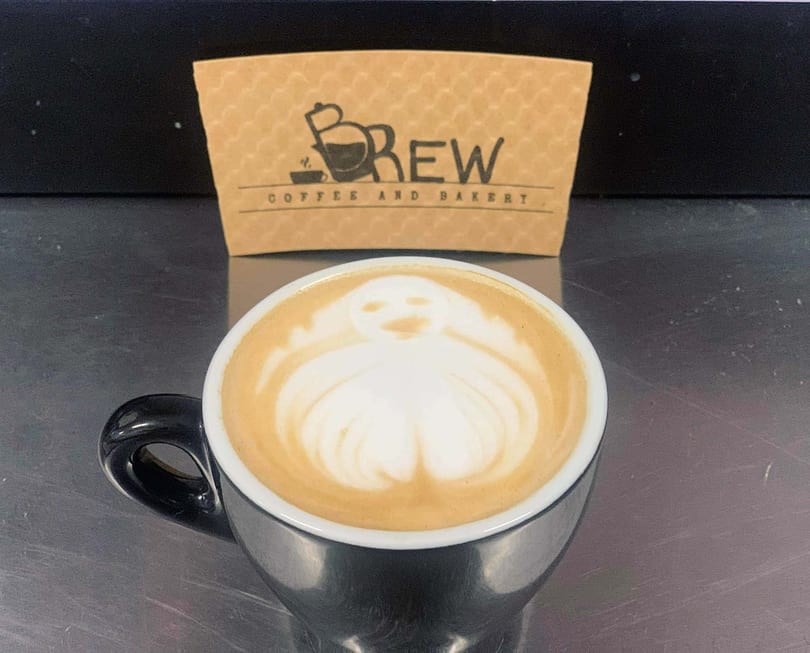 A black coffee cup with latte art depicting a ghost, with a "Brew Coffee and Bakery" sleeve behind it.