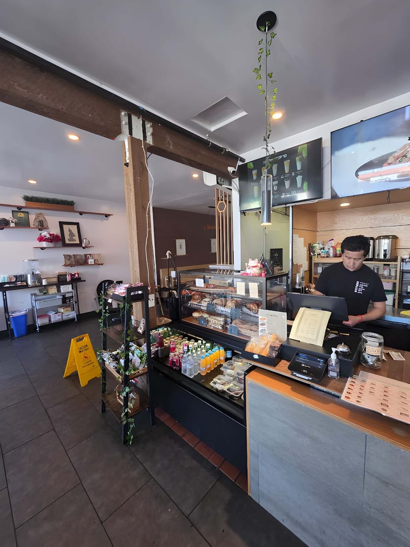 A man stands behind a counter with pastries, drinks, and a cash register in a cafe.