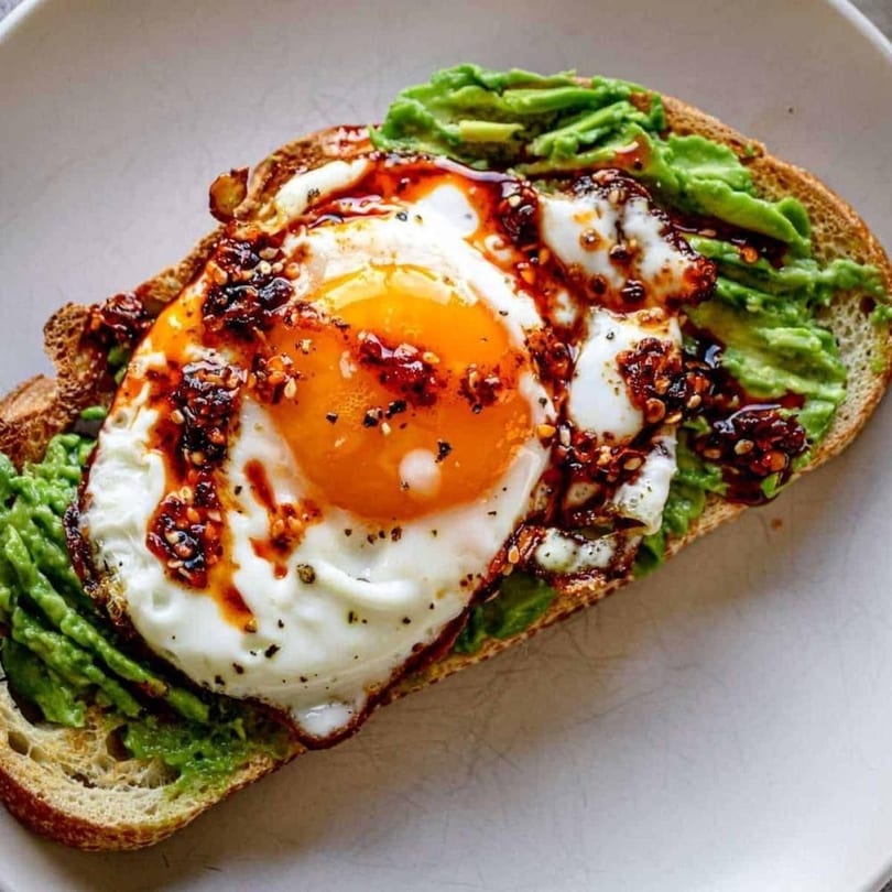 Avocado toast topped with a fried egg and chili oil on a white plate.