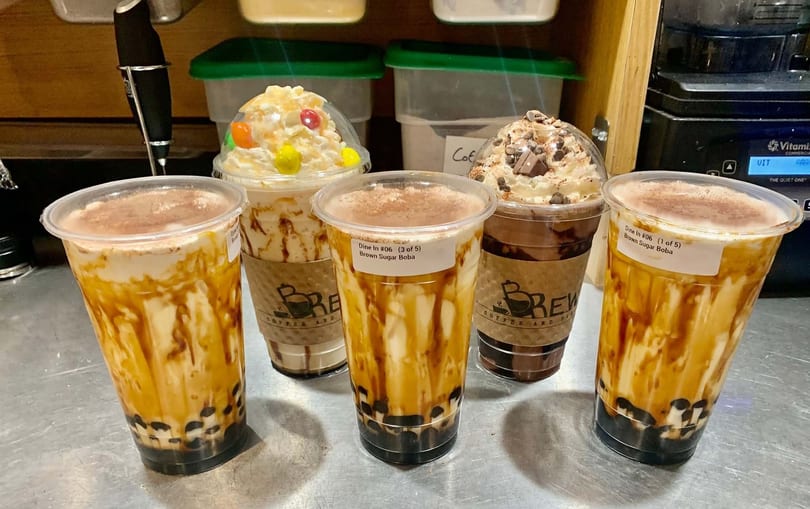 Five boba drinks, three brown sugar boba, one with candy, and one chocolate, on a metal counter.