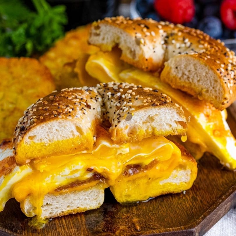 Two everything bagels overflowing with melted cheese and egg, served on a wooden board.