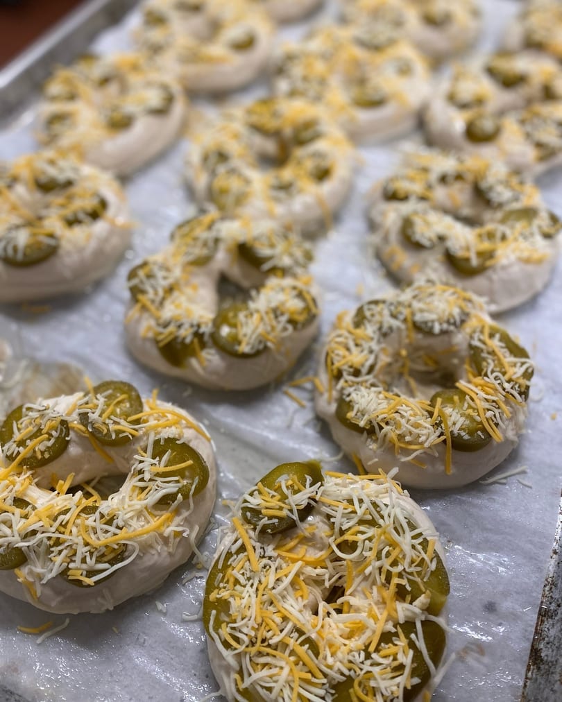 Unbaked bagels topped with sliced jalapeños and shredded cheese on a baking sheet.