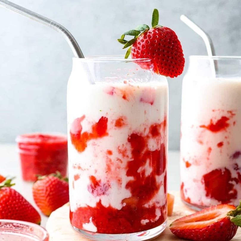 Two glasses of strawberry milk with fresh strawberries and metal straws on a light background.
