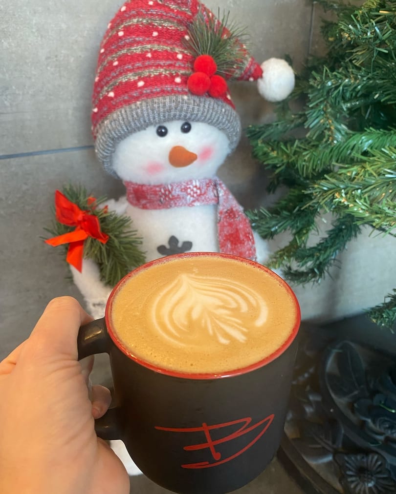 Hand holding a black mug with latte art, next to a snowman decoration and a Christmas tree.